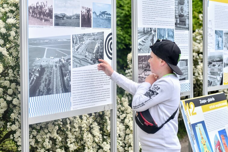 Zdjęcie. Plener. Chłopiec wskazuje palcem na plansze wystawy. 