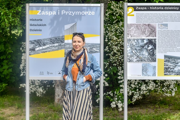 Zdjęcie. Plener. Na zdjęciu Katarzyna Kurkowska na tle wystawy plenerowej Historie Gdańskich Dzielnic. 
