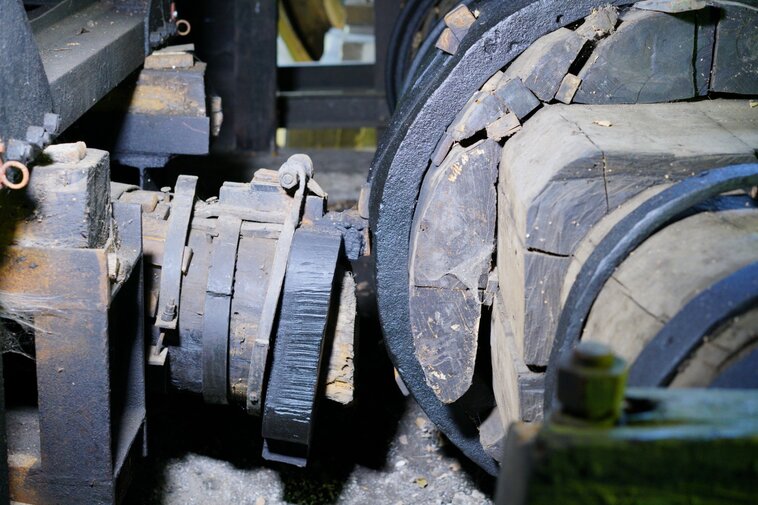 Zdjęcie przedstawia zbliżenie na elementy konstrukcji drewnianego młota wodnego znajdującego się we wnętrzu kuźni. Widoczne metalowe okucia łączące drewniane elementy. 