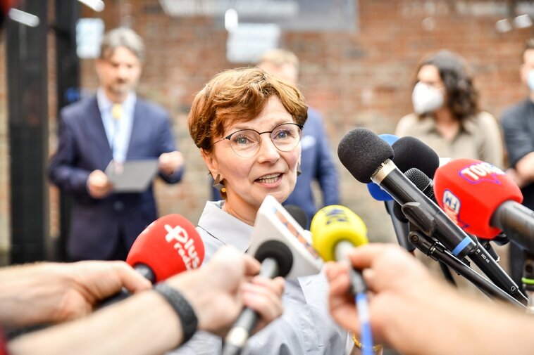 Zdjęcie. Renata Adamowicz. Wokół mikrofony. W tle ceglany mur i ludzie. 