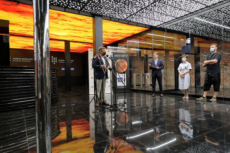 Wnętrza Wielkiego Młyna. Przemawia Waldemar Ossowski, dyrektor Muzeum Gdańska. W tle od lewej Andrzej Gierszewski, Renata Adamowicz oraz Robert Sochacki. W tle pomieszczenie w odcieniach czerni i bursztynu.  