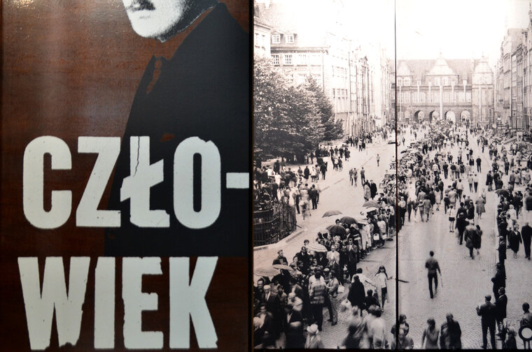 Dwie plansze. Duży napis Człowiek. Obok zdjęcie Długiego Targu. Kolejka osób do Dworu Artusa, chcących oddać honory przy urnie ze szczątkami Majora.