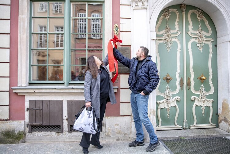 Zdjęcie. Anna Kriegseisen (z lewej) oraz Bartosz Nawacki (z prawej) rozwiązują wstęgę wokół uchwytu repliki dzwonka. W tle fasada Domu Uphagena.