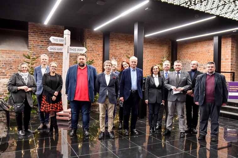 Zdjęcie. Wnętrza Muzeum Bursztynu. Od lewej stoją: dr Ewa Szymańska, dr Janusz Marszalec, Ewa Joszczak, Robert Pytlos, prof. Krzysztof Polkowski, prof. Piotr Niemcewicz, dr Sylwia Bykowska (oboje w drugim rzędzie), dr Zbigniew Canowiecki, Barbara Frydrych, Tomasz Wierzchowski, dr hab. Waldemar Ossowski, prof. Edmund Kizik i prof. Igor Hałagida.  