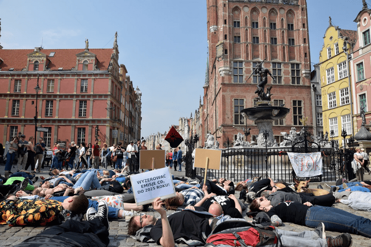 Zdjęcie. Kobiety i mężczyźni leżący przed Fontanną Neptuna w Gdańsku. Widoczny transparent z napisem: wyzerować emisję CO2 do 2025 roku. 