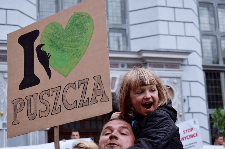 Zdjęcie. Mężczyzna z małą dziewczynką na plecach. Nad nimi transparent z napisem "I Love Puszcza" i sercem. W tle fragment fasady Dworu Artusa. 