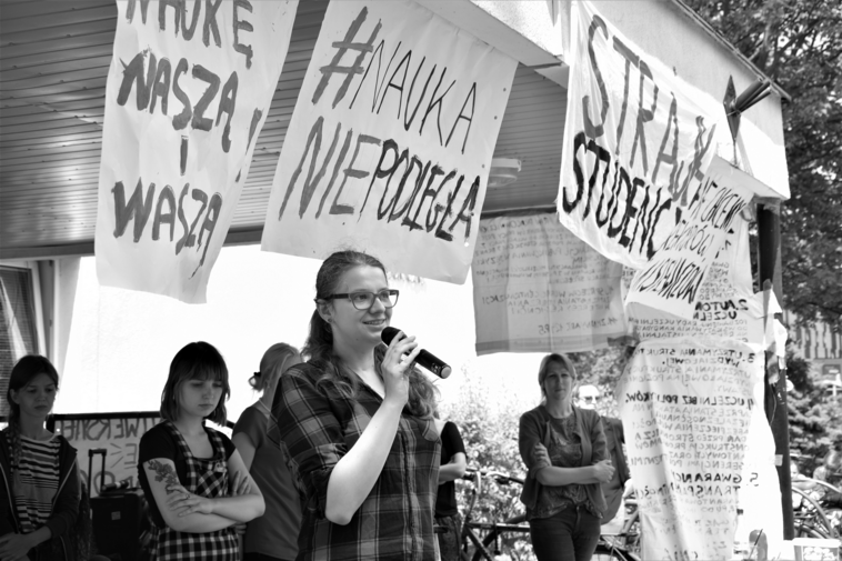 Zdjęcie czarno-białe. Gdański protest akademicki w 2018 roku. Na scenie przemawiają studentki. Nad nimi 3 transparenty z napisami "Za naukę naszą i waszą", "Strajk studencki" oraz "#naukaniepodległa". 