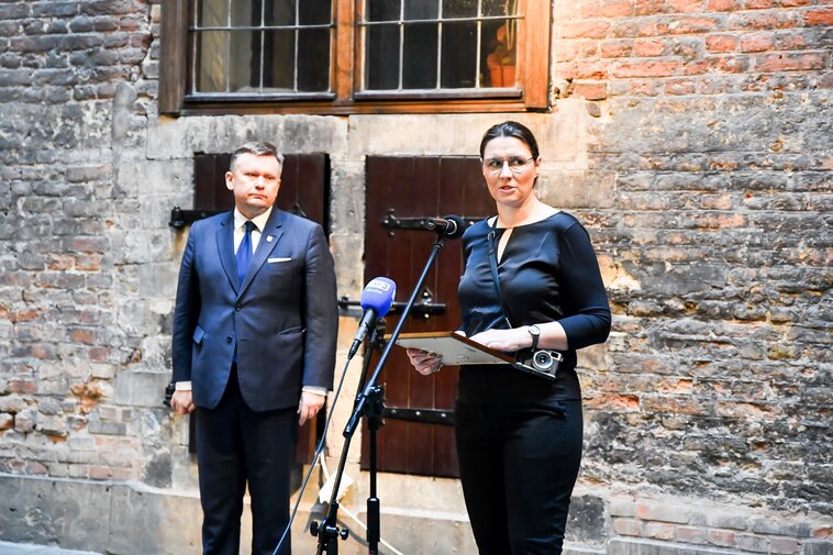 Zdjęcie. Waldemar Ossowski (z lewej) i przemawiająca Karolina Misztal (z prawej). W tle fragment gotyckiego muru z okiennicami.  