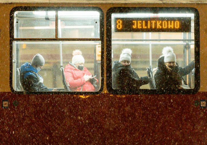 Zdjęcie. Fragment tramwaju z dwoma oknami. Wewnątrz siedzą 4 osoby z maseczkami zakrywającymi częściowo twarz. Patrzą  w telefon, albo w szybę (w kierunki widza). W prawym oknie u góry napis "8 Jelitkowo". Pada śnieg.   