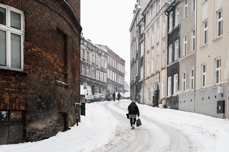 Zdjęcie. Zaśnieżona ulica Biskupia i jej kamienice. Pod górkę wchodzi gdańszczanka niosąca torby z zakupami. 