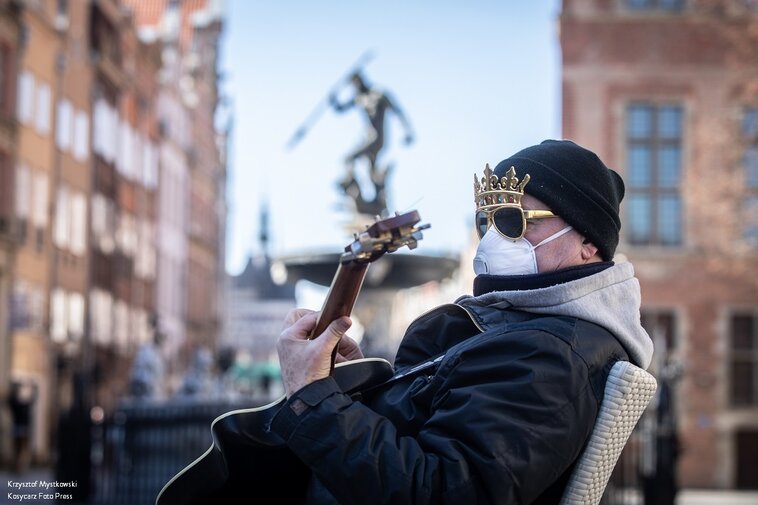 Zdjęcie. Mężczyzna z maseczką na twarzy i koroną na głowie gra na gitarze. W tle Fontanna Neptuna. 