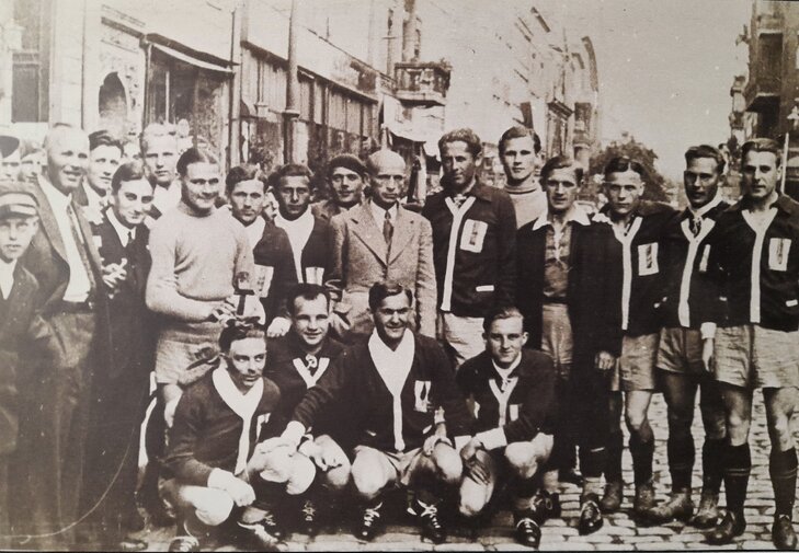Czarno-białe zdjęcie z ok. 1932 roku. Członkowie sekcji piłkarskiej wraz z władzami klubu i sympatykami pozują do zdjęcia wśród toruńskich kamienic. 