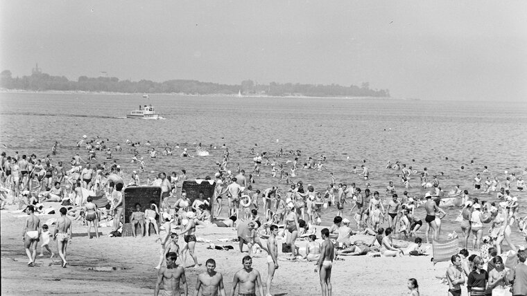 Fragment czarno-białej fotografii. Na zdjęciu plaża na gdańskich Stogach. Na brzegu i w wodzie liczni ludzie. W tle fragment brzegowy w kierunku Jelitkowa. Na morzu niewielki statek wycieczkowy. 