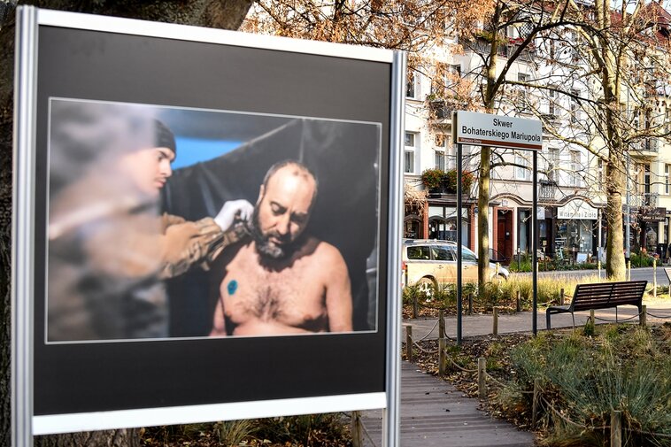 Wystawa plenerowa. W kadrze żołnierz podczas golenia. W tle otoczenie Skweru Bohaterskiego Mariupola. Budynki i drzewa.  