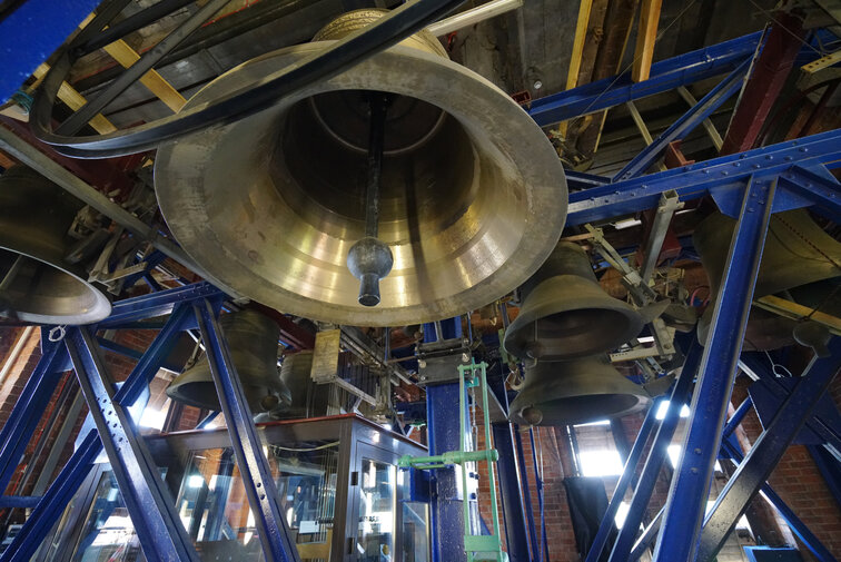 Zdjęcie wykonane w wieży kościoła św. Katarzyny (Muzeum Nauki Gdańskiej). Widok na zespół dzwonów – carillon, zawieszony na stalowej konstrukcji. Na pierwszym planie widoczny od dołu dzwon. W dali widoczne cztery dzwony oraz przeszklone niewielkie pomieszczenie, w którym gra carillonista. 