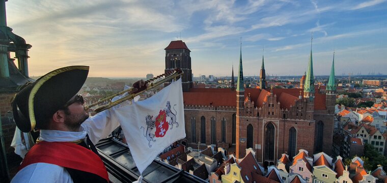 Zdjęcie przedstawia trębacza ubranego w historyczny strój. Do trąbki przymocowany chwytak, na nim flaga a na niej z lwy trzymające herb Gdańska. W tle zabudowania oraz Bazylika Mariacka.  
