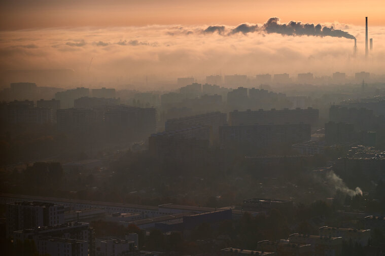 Panorama osiedla Zaspa. Widok z góry na osiedle bloków. W prawym rogu widać kominy, z których unosi się smuga dymu, aż nad bloki. Światło jest ciepłe, ale lekko przygasa, jak podczas wschodu lub zachodu słońca.