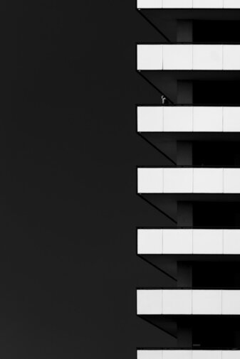 Zdjęcie czarno białe. Nowoczesny budynek. Prawa część zdjęcia jest cała czarna, po prawej widać białe poziome pasy, płyty osłaniające balkony. Na jednym z nich widać sylwetkę człowieka, słabo wyraźną.