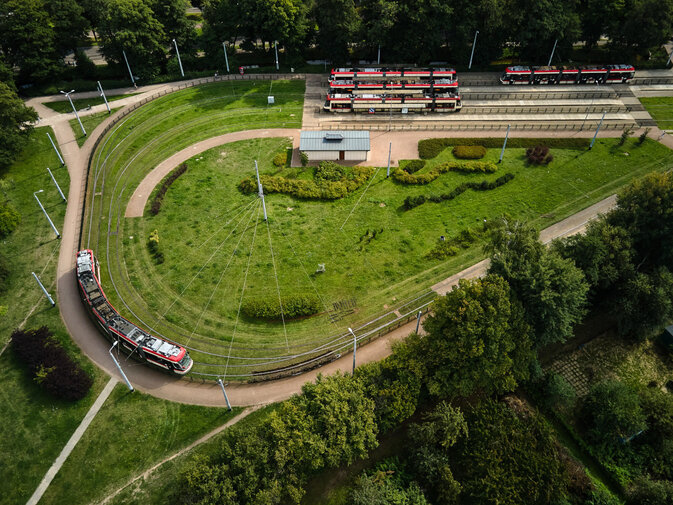 Zdjęcie. Widok z lotu ptaka na pętlę tramwajową. Tory zakręcają, tworząc kształt w kształcie łzy. Pomiędzy nimi rośnie trawa, a wokół drzewa. Przy przystanku stoją 3 tramwaje, jeden zawraca na pętli.