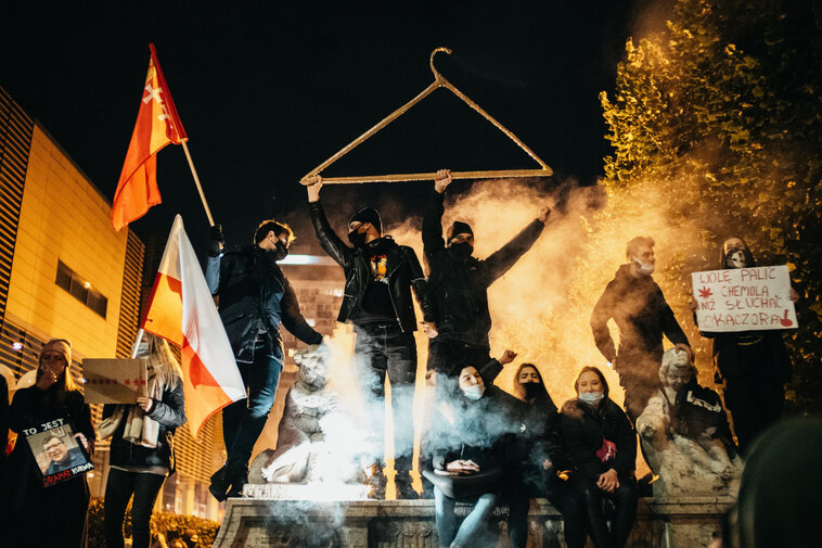 Zdjęcie wieczorne z protestu. 10 osób w maseczkach ochronnych stoi, trzymają transparenty z hasłami przeciw partii rządzącej i zaostrzeniu prawa aborcyjnego. Dwaj mężczyźni pośrodku trzymają w górze wielki wieszak. Za nimi unosi się smuga dymu.