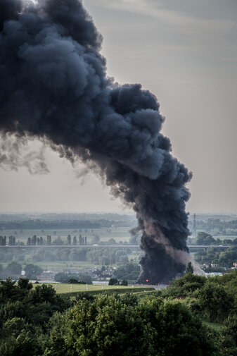 Zdjęcie. Widok z oddali na ogromny słup czarnego dymu, unoszący się ponad miastem. Na pierwszym planie widać drzewa, a w oddali pola, drzewa i nieliczne zabudowania.