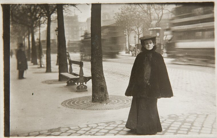 Olga Boznańska na ulicy miasta. W tle ulica, drzewa i ławki. 