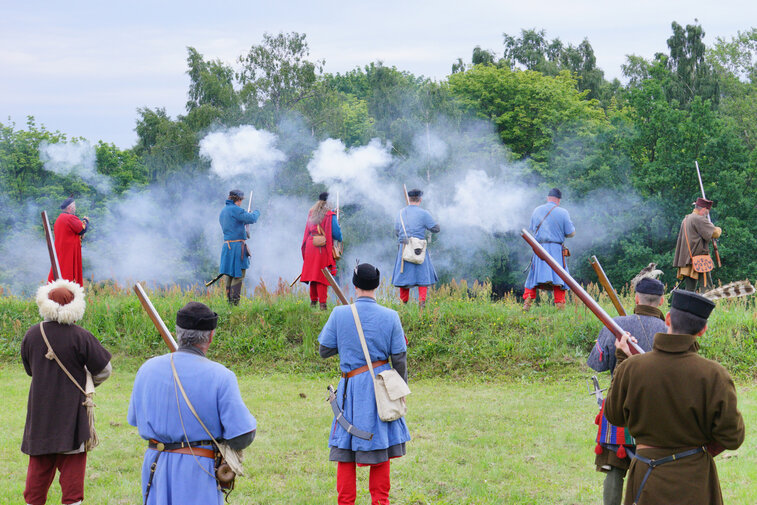 Zdjęcie przedstawia rekonstrukcję walk w Twierdzy Wisłoujście podczas festiwalu rekonstrukcyjnego. Widać jedenaście postaci w historycznych strojach strzelających z muszkietów.