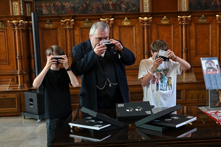 Zdjęcie. Dwór Artusa, Wielka Hala. Dwaj chłopcy oglądają zdjęcia przez okulary stereoskopowe. Pomiędzy nimi stoi mężczyzna w garniturze stylizowanym na 19 wiek, też ogląda zdjęcia przez okulary.