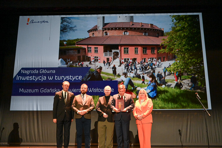 Od lewej Mieczysław Struk - Marszałek Województwa Pomorskiego, Maciej Flis - kierownik Twierdzy Wisłoujście, Tomasz Wierzchowski - z-ca dyrektora Muzeum Gdańska, Waldemar Ossowski - dyrektor Muzeum Gdańska oraz Marta Chełkowska - dyrektorka Departamentu Turystyki i Sportu Urzędu Marszałkowskiego Województwa Pomorskiego i prezeska Pomorskiej Regionalnej Organizacji Turystycznej. W tle zdjęcie lotnicze Twierdzy Wisłoujście na Martwą Wisłą. 