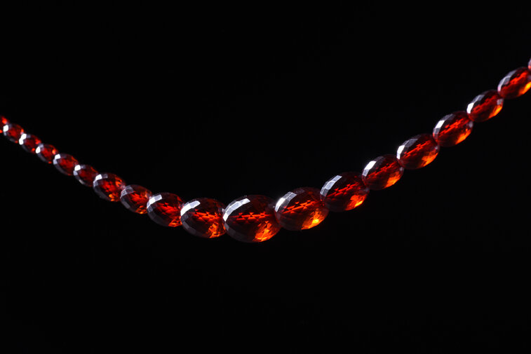 Sznur korali fasetowanych. Paciorki owalne o różnej wielkości, zwiększające się od zapięcia do środka sznura. Zapięcie plastikowe, dwuczęściowe, zamykane przez zakręcanie. Paciorki przejrzyste w kolorze koniakowym - imitacja bursztynu.