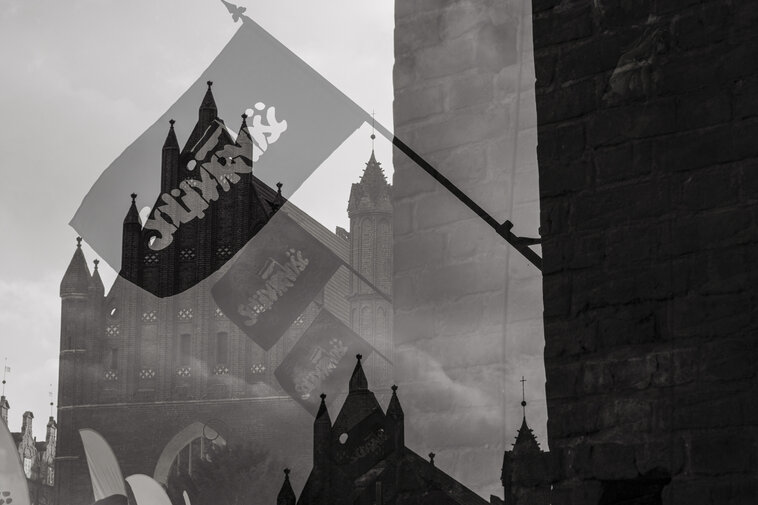 Czarno-biała scena z Bazyliką św. Brygidy. Flagi „Solidarności” powiewają na wietrze przed masywną, gotycką bryłą kościoła.