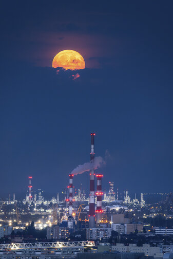 Pełnia księżyca nad nocnym Gdańskiem. Jasny, pomarańczowy księżyc wznosi się nad kominami rafinerii i rozświetloną panoramą miasta.