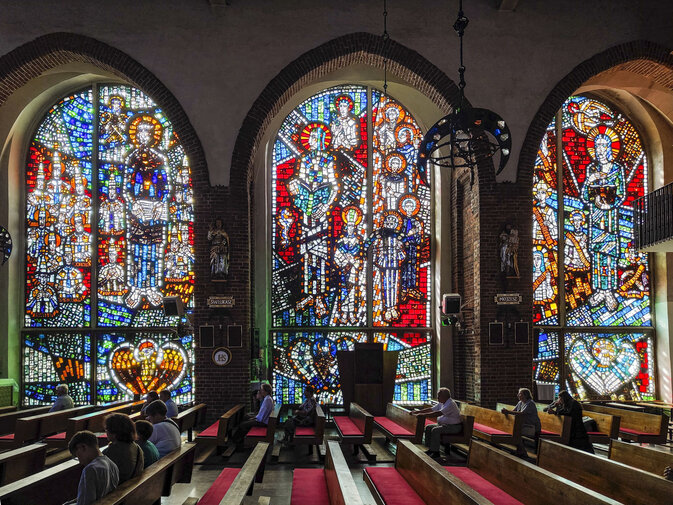 Wnętrze kościoła św. Barbary w Gdańsku. Wysokie, kolorowe witraże wypełniają przestrzeń światłem, nad ławkami siedzą wierni.