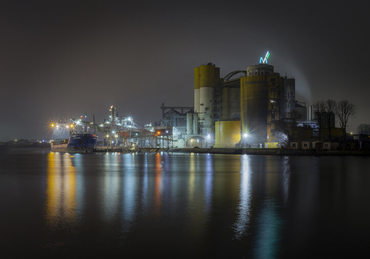 Nocny widok na terminal w Gdańsku. Oświetlone silosy i infrastruktura portowa odbijają się w spokojnej tafli wody, tworząc industrialny pejzaż po zmroku.