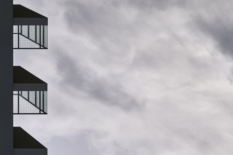 Fragment nowoczesnego budynku na Morenie. Minimalistyczna elewacja z metalowymi balkonami na tle pochmurnego, jasnoszarego nieba.