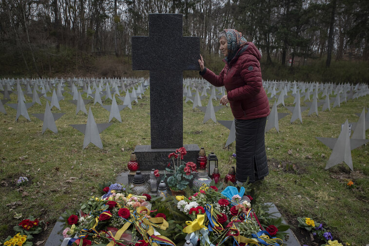 Starsza kobieta w czerwonej kurtce stoi przy granitowym krzyżu, dotykając nagrobka. U stóp krzyża kwiaty i znicze; w tle rzędy metalowych gwiazd na wojskowym cmentarzu.