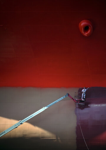 Pracownik na podnośniku maluje ogromny kadłub statku. Górna część ściany intensywnie czerwona, niżej ciemniejszy pas; skala podkreślona małą sylwetką człowieka.

