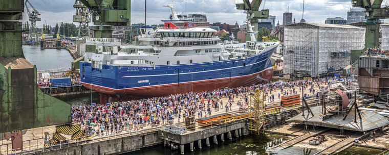 Tłum ludzi zgromadzony przy wodowaniu dużego niebiesko-białego statku w stoczni. Wokół dźwigi portowe, nabrzeże i zabudowa przemysłowa.