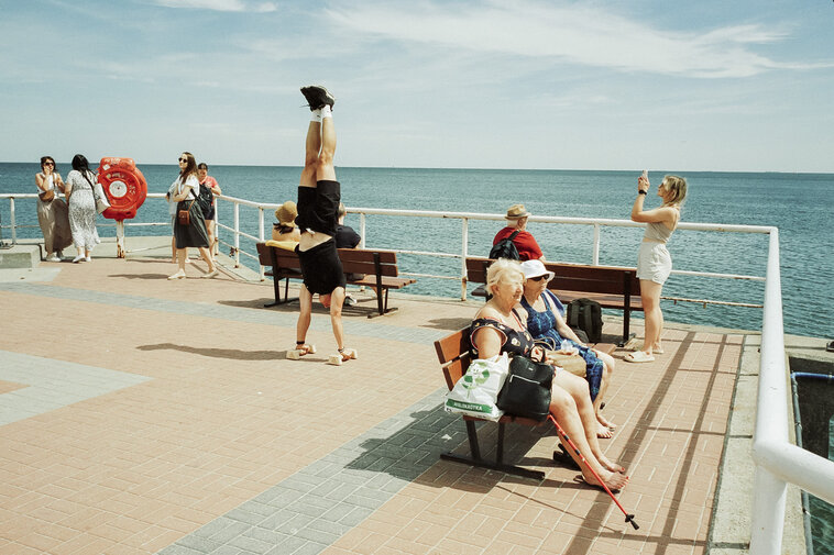 Letnia scena na molo: mężczyzna stoi na rękach, inni spacerują i siedzą na ławkach. W tle spokojne morze i bezchmurne niebo.