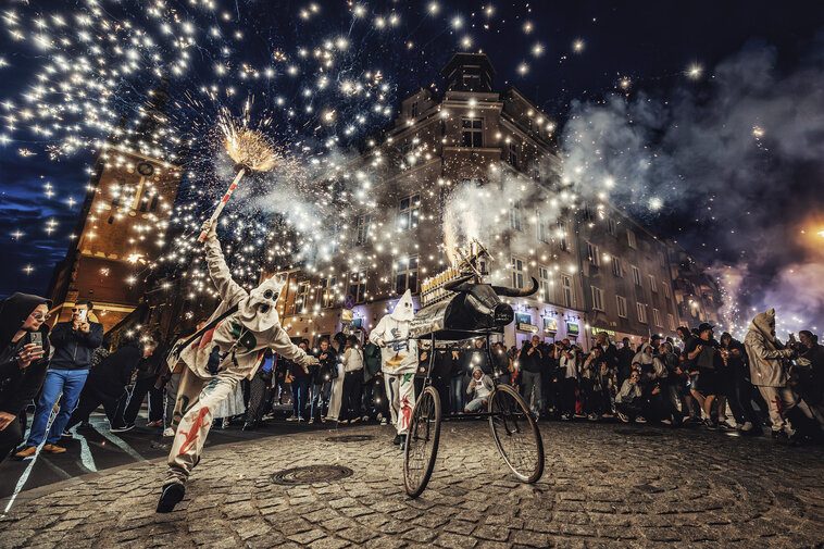 Nocny pokaz uliczny z ogniem: performer w kostiumie rozpala iskry nad głową, obok wózek artystyczny. Wokół zgromadzona publiczność na staromiejskiej ulicy.