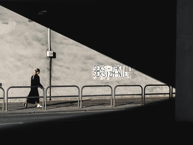 Kobieta ubrana na czarno idzie chodnikiem pod wiaduktem. Na szarej ścianie napis „SEKS – TAK, SEKSIZM – NIE”, przecięty ostrym pasem światła i cienia.