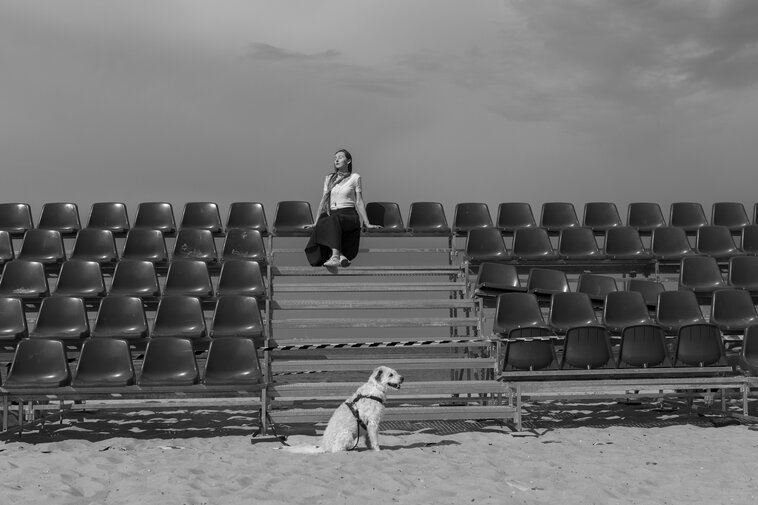 Kobieta siedzi samotnie na środku pustych trybun ustawionych na plaży. Na piasku przed nią siedzi pies; całość w czerni i bieli, z szerokim niebem w tle.