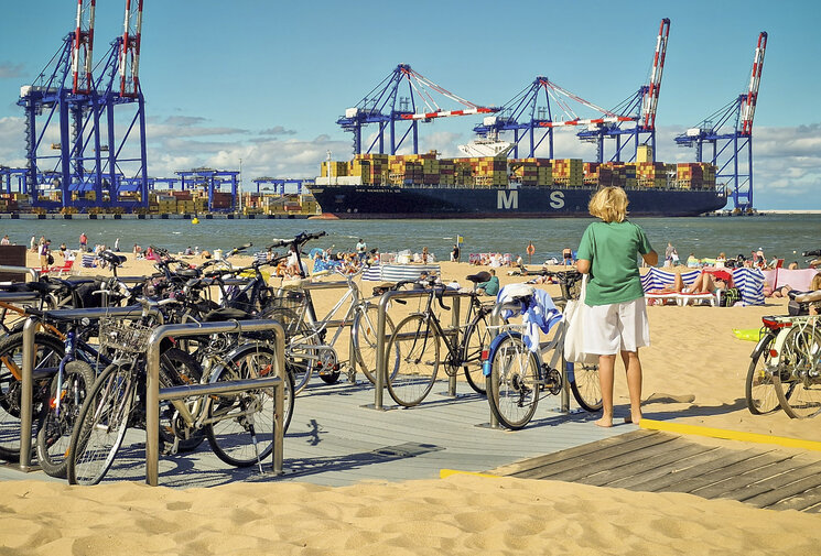 Kobieta stoi boso na plaży obok zaparkowanych rowerów i patrzy na ogromny kontenerowiec oraz suwnice portowe; plażowicze wypoczywają na piasku.