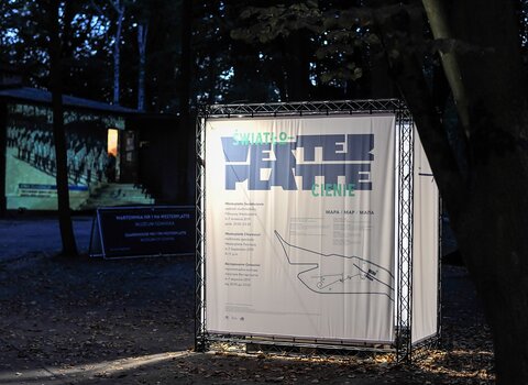 Zdjęcie przedstawia kubik informacyjny festiwalu "Westerplatte. Światłocienie". Bryła stoi na tle lasu. W tle prostopadłościenna bryła Wartowni nr 1 na Westerplatte. Na jej ścianie frontowej wyświetlany jest film.  