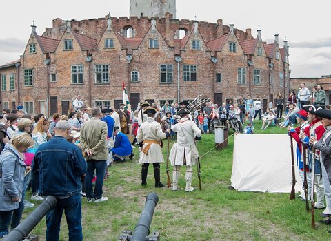 Majówka w Twierdzy Wisłoujście. Na dziedzińcu stoją ludzie, pośród nich rekonstruktorzy przebrani w stroje żołnierzy szykujący się do opalenia muszkietów. W tle kamieniczki wieńca i szczyt wieży. 