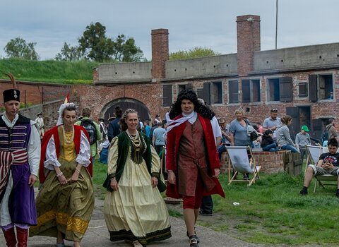 Majówka w Twierdzy Wisłoujście. Parada maszerujących przez plac rekonstruktorów i rekonstruktorek. Po prawej rząd turystów. W tle zabudowania koszar i wały. 