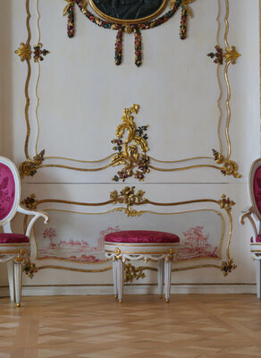 Zdjęcie przedstawia wnętrza Domu Uphagena. W centrum biała ściana, na prawo i lewo od niej szkarłatne, zdobione w formie smukłych ośmiokątów tapety. Na drewnianej podłodze stoją dwa białe krzesła obite szkarłatną tkaniną w kolorze tapet. Pomiędzy nimi niskie siedzisko obite tkaniną w tym samym kolorze.   
