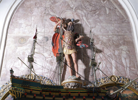 Zdjęcie przedstawia model okrętu zwisający na linkach. Z nim rzeźba św. Krzysztofa na tle imitacji oryginalnego obrazu, który spłonął podczas bombardowań pod koniec II wojny światowej. U góry poprzeczka, do której za pomocą linek zamocowany jest okręt.