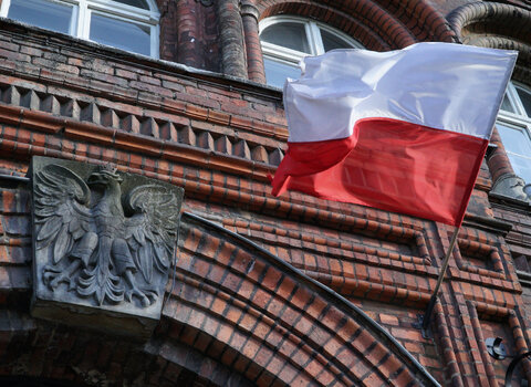 Zdjęcie przedstawia fragment fasady gmachu Poczty Polskiej. Nad drzwiami godło RP. Po prawej łopocze flaga Polski. 