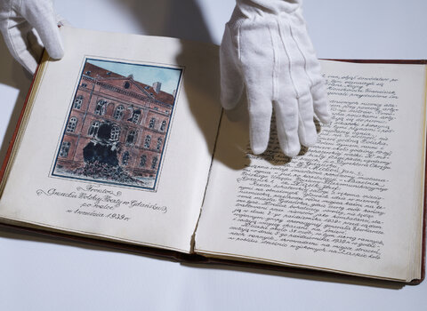 Zdjęcie przedstawia pamiętnik / książkę, którą trzymają donie w rękawiczkach. Na lewej stornie rysunek przedstawiający uszkodzony budynek z wyrwą powstałą wskutek niemieckiego ostrzału. Na prawej ręczne, kaligrafowane pismo.  